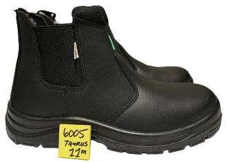A pair of black Chelsea-style safety boots with elastic side gore panels and a thick lug sole, photographed on a light surface. A small yellow handwritten price tag is placed in front of the boot.